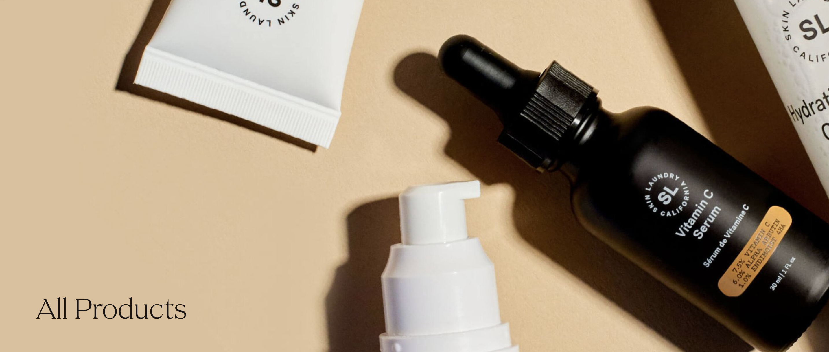 A flat lay of various Skin Laundry skincare products, including a Vitamin C Serum in a black dropper bottle, a white pump bottle, and a white tube, all arranged on a light beige background with text "All Products."