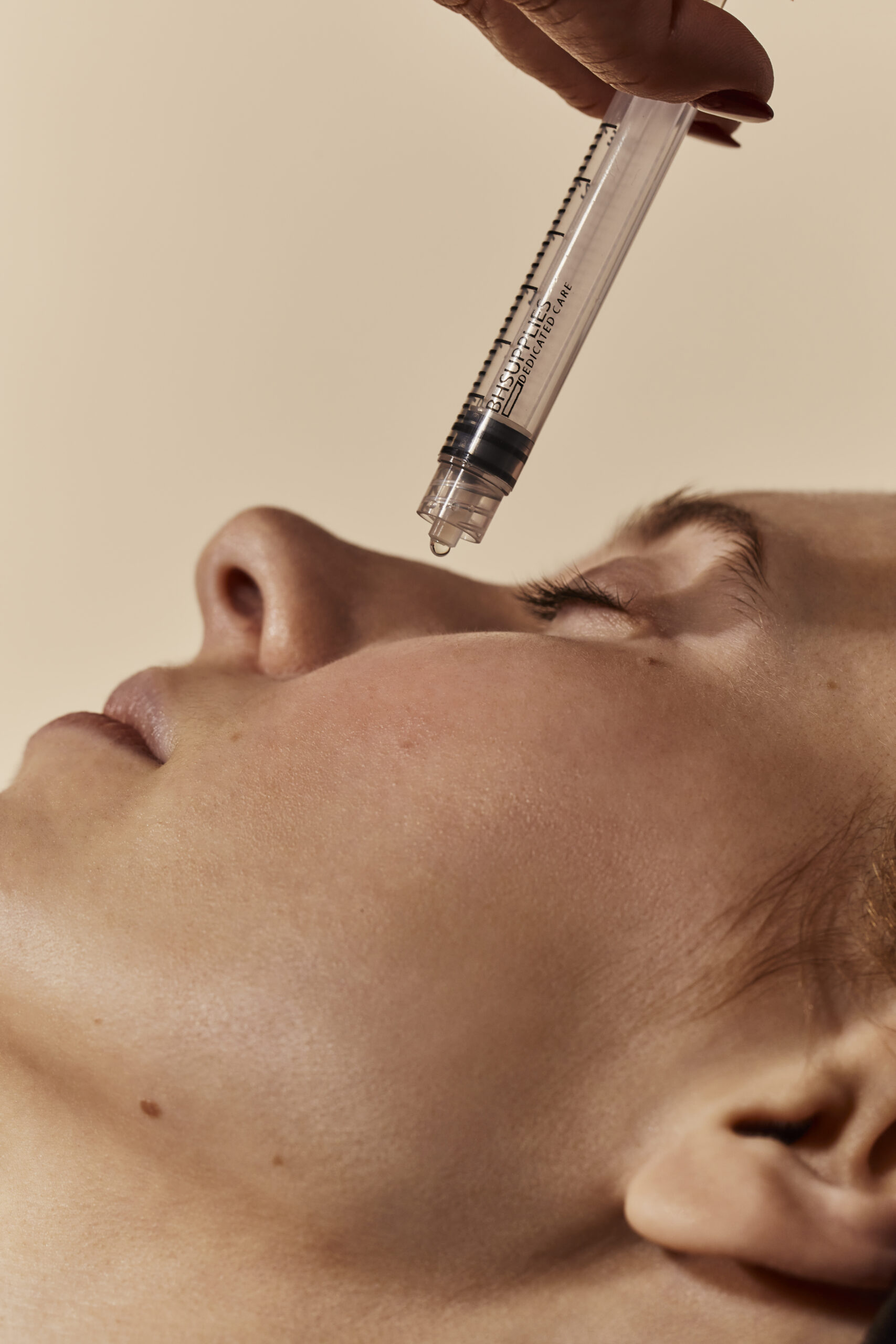 A close-up of a person's face with closed eyes, receiving a topical application of serum from a syringe-like applicator on their forehead, suggesting a precise and potent skincare treatment.