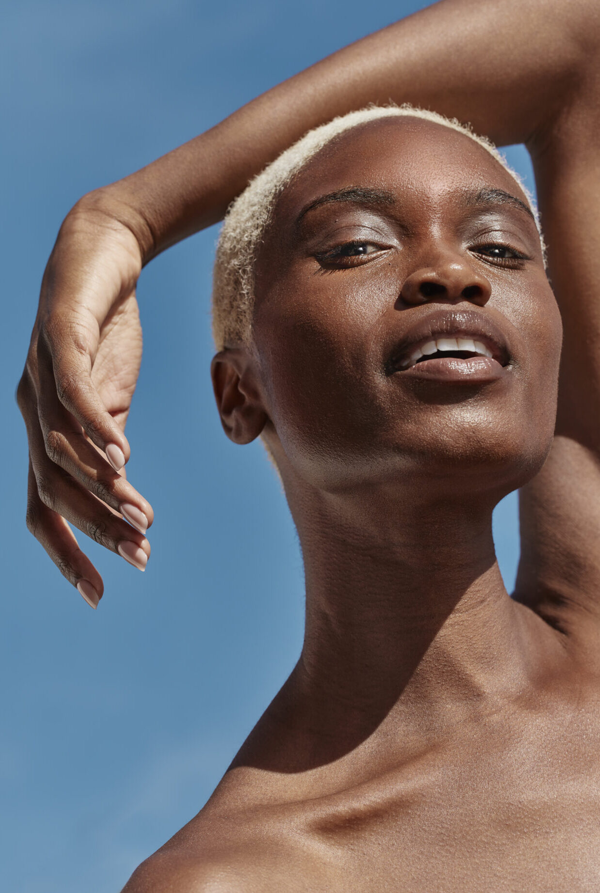 A radiant person with short, light-colored hair and dark, dewy skin, raising one arm over their head and looking upwards with a confident expression against a bright blue sky, highlighting clear skin.