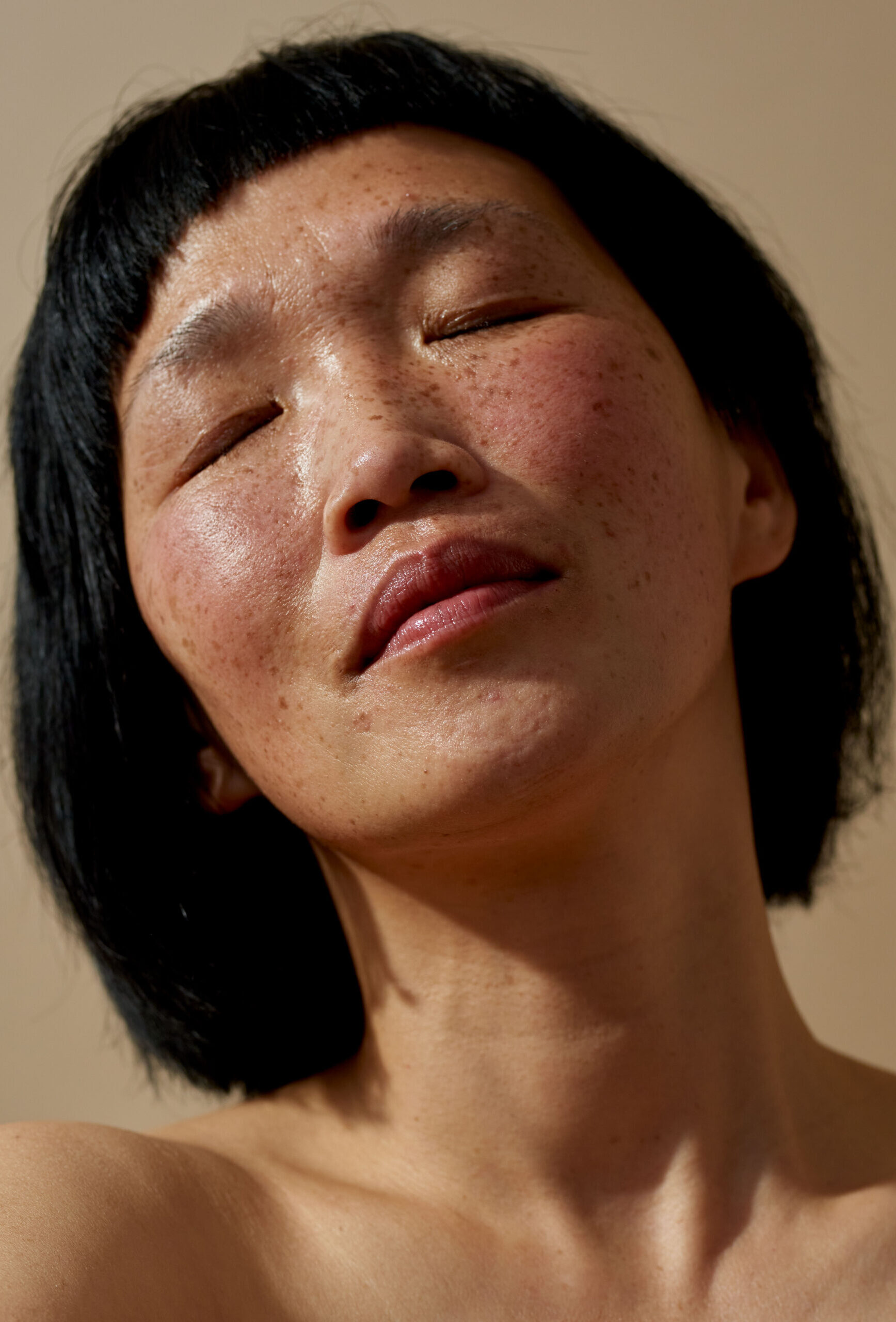 A close-up portrait of a person with short dark hair and freckles, with their eyes gently closed and head slightly tilted back, conveying a sense of calm and natural beauty.