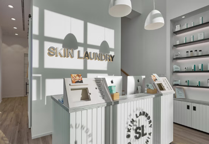 The bright and modern reception area of a Skin Laundry clinic, featuring a white slatted desk with digital check-in stands, computers, and a prominent "SKIN LAUNDRY" sign on the wall. Shelves with skincare products are visible on the right.