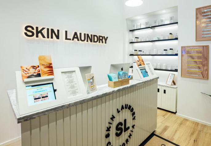 The reception desk area of a Skin Laundry clinic, featuring a white slatted desk with a marble top, digital screens for check-in or information, product displays, and illuminated "SKIN LAUNDRY" lettering on the wall.