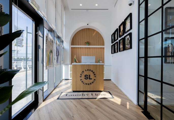 The bright and modern reception area of a Skin Laundry clinic, featuring a wooden slatted desk with the brand's circular logo, a "Laundry I Love" doormat, an arched alcove, and large windows bringing in natural light.