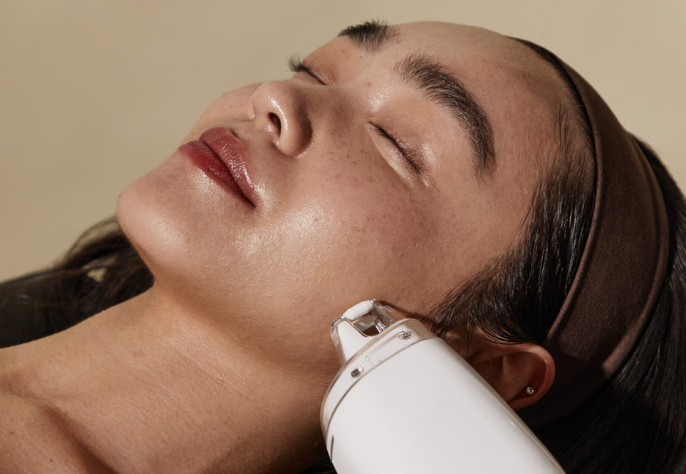 A person with closed eyes receiving a facial treatment with a white handheld device being glided along their jawline, suggesting a cleansing or massage procedure.