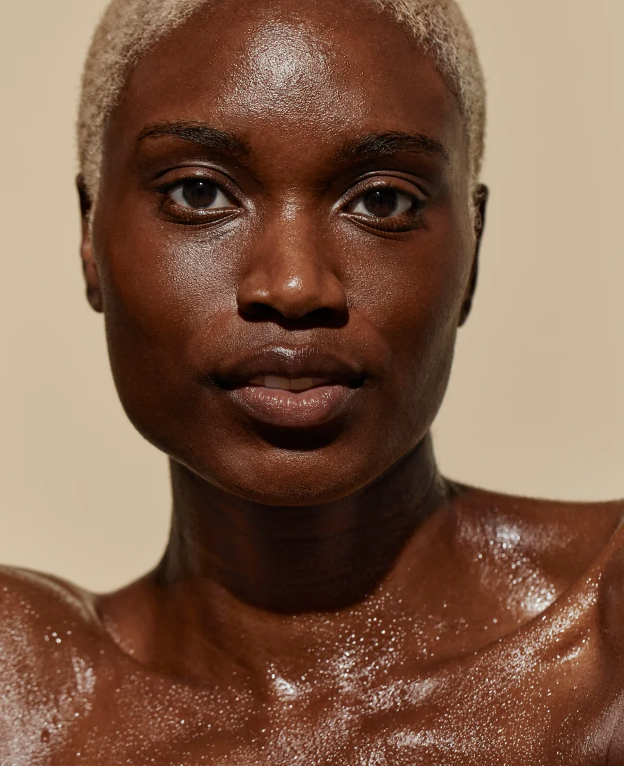 A close-up portrait of a person with dark, radiant skin and short light-colored hair, looking directly at the viewer with a confident expression, showcasing a dewy, hydrated complexion.