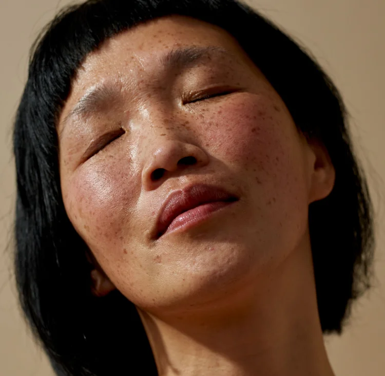 A close-up portrait of a person with short dark hair and freckles, with their eyes gently closed and head slightly tilted back, conveying a sense of calm and natural beauty.
