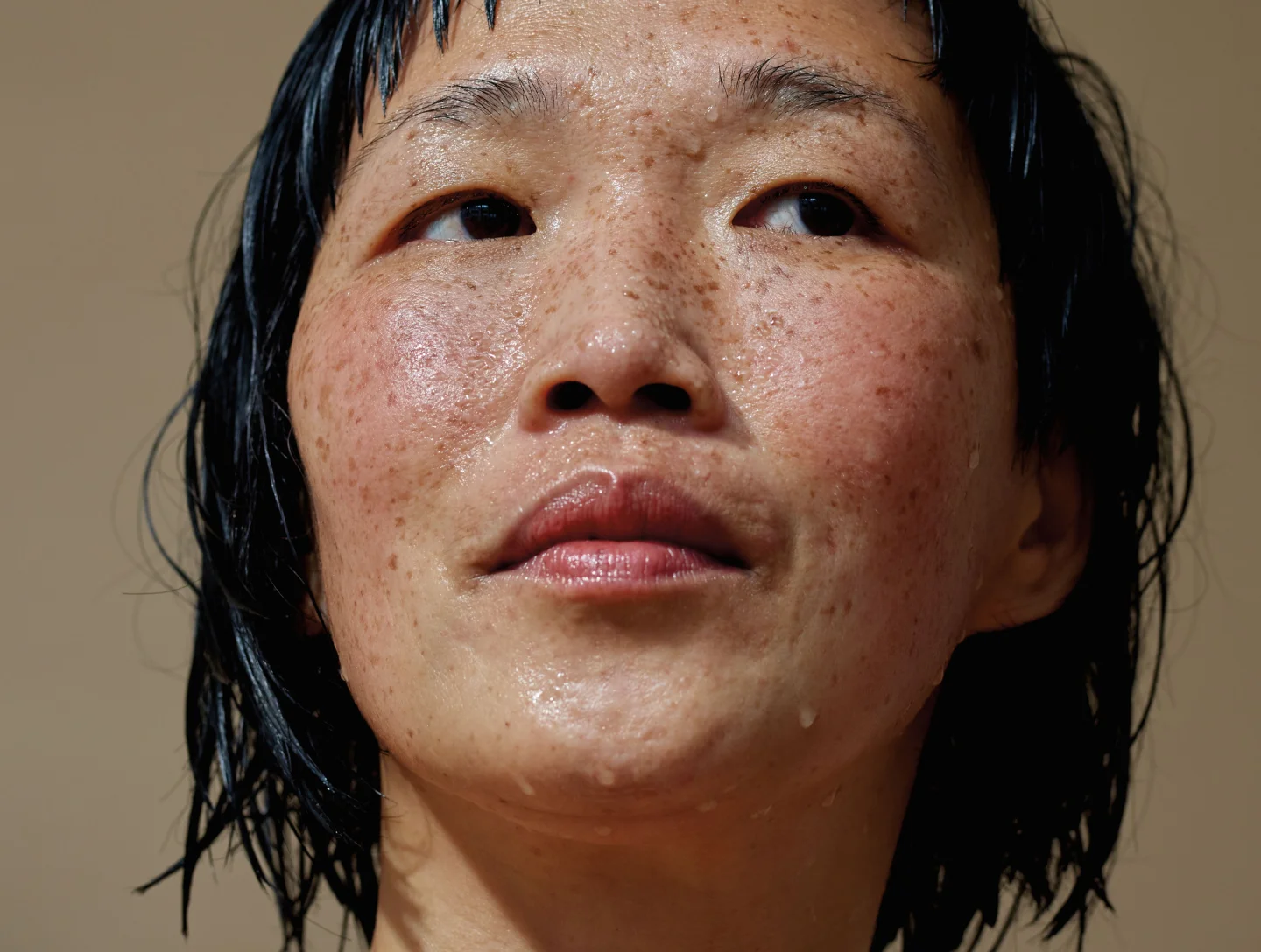 A close-up portrait of a person with freckles and wet, glistening skin, looking upwards, suggesting a state of intense hydration or post-treatment glow.