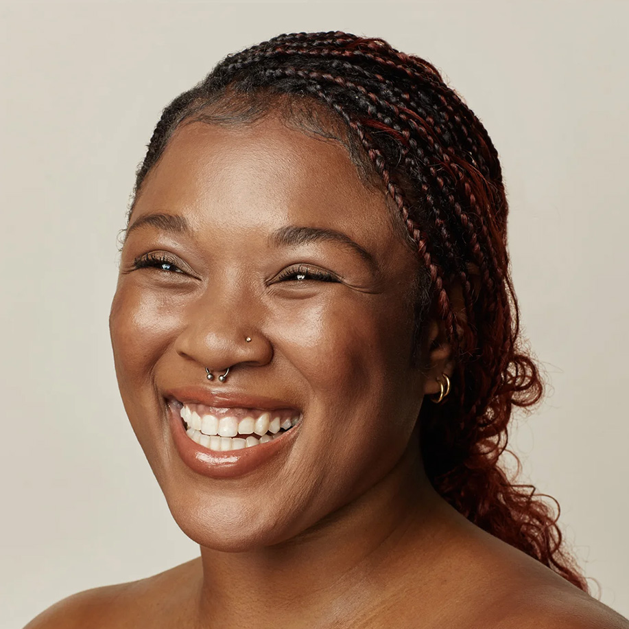Smiling person with radiant, clear skin and dark braided hair, likely after a skin treatment, showcasing healthy glow. Features nose piercing and hoop earrings.