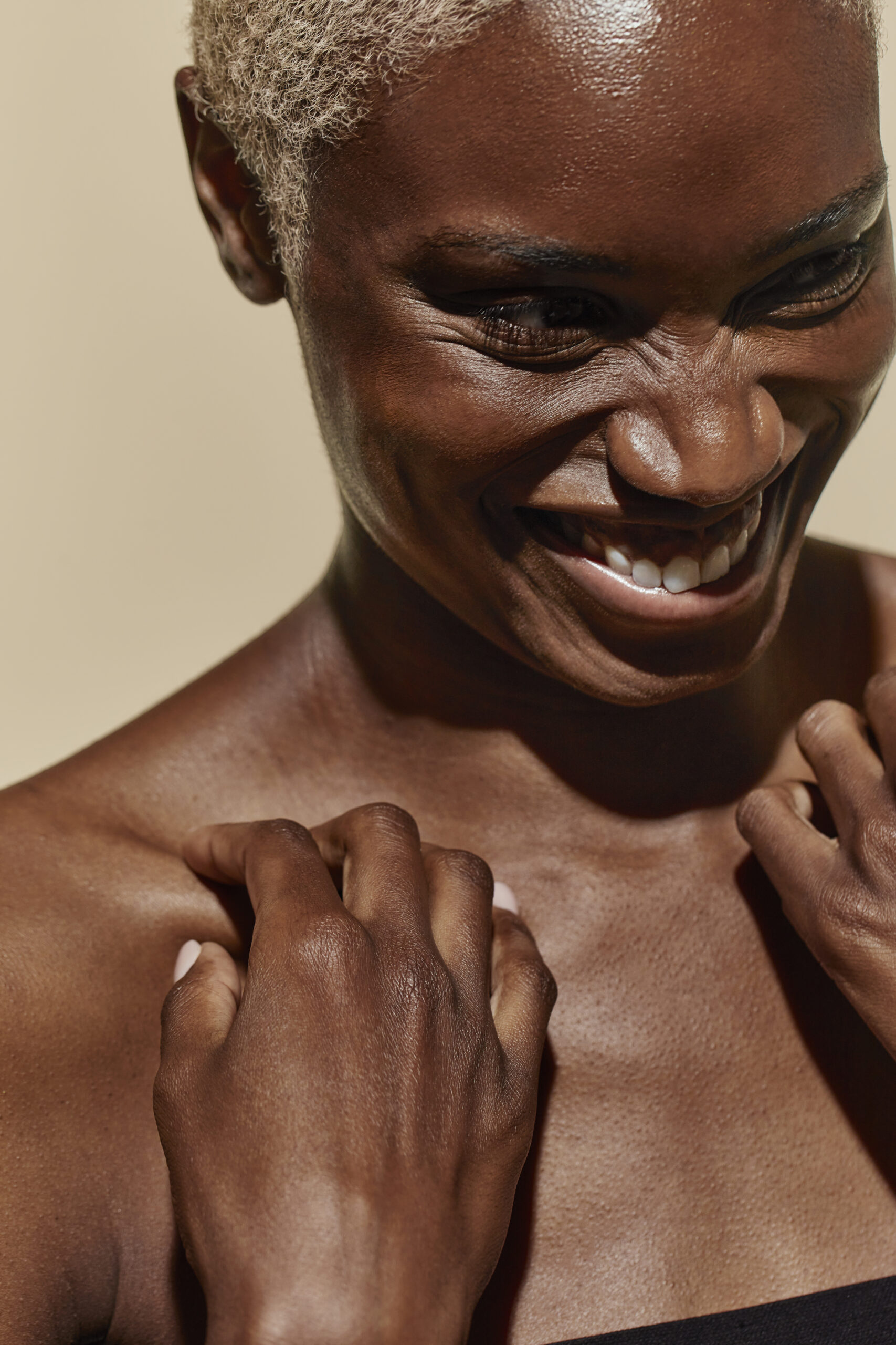 A close-up portrait of a person with radiant dark skin and short light-colored hair, smiling broadly with closed eyes and visible joy, touching their shoulders.