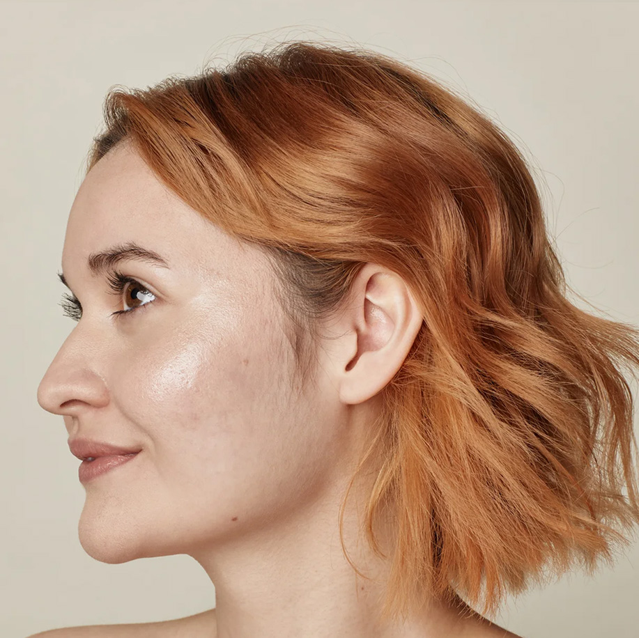 Side profile of a person with fair, glowing skin and short, wavy reddish-blonde hair, showing a healthy, hydrated complexion. Highlights skin texture and luminosity.