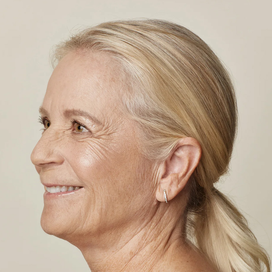 Side profile of a smiling person with clear, healthy skin and blonde hair tied back, wearing a small hoop earring. Highlights natural skin texture and glow.