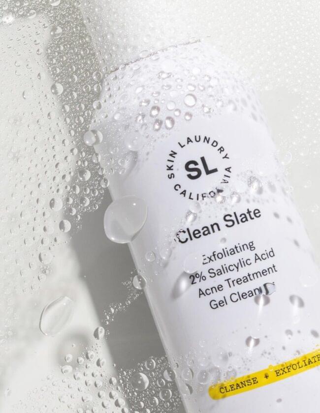 A white bottle of Skin Laundry "Clean Slate" Exfoliating 2% Salicylic Acid Acne Treatment Gel Cleanser, covered in water droplets, with a yellow bar indicating "CLEANSE + EXFOLIATE."