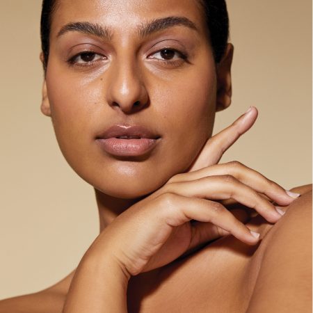 A close-up portrait of a woman with glowing, warm-toned skin and her hair pulled back, looking directly at the camera with one hand gently touching her shoulder, highlighting her clear and radiant complexion.